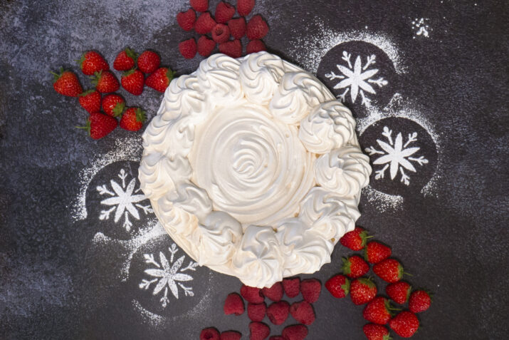 An empty pavlova case surrounded by fresh strawberries, raspberries and icing sugar snowflakes